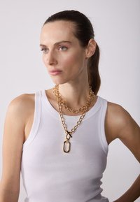 Goldene Kettenhalskette mit mehreren Gliedern und einem auffälligen übergroßen Verschluss, getragen über einem weißen gerippten Tanktop.
