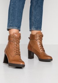 Bottes en cuir marron à la cheville avec un design à lacets, des accents en métal doré, et un talon bloc texturé, portées avec un jean en denim retroussé.