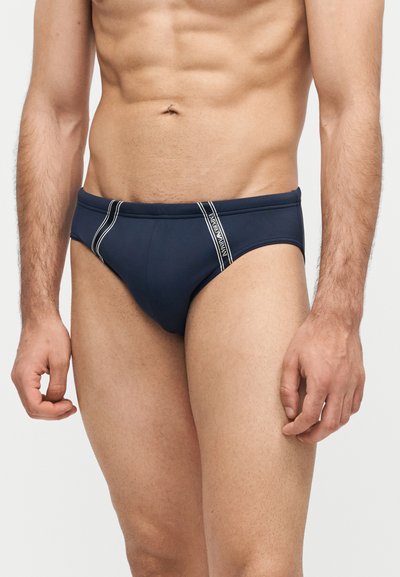 Άνδρας που φοράει μαγιό navy blue σε στυλ σλιπ με λευκές ριγέ λεπτομέρειες και λογότυπο Emporio Armani στο μπροστινό αριστερό μέρος.