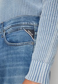 Jasnoniebieski dziergany sweter z prążkowanym mankietem i wyblakłe niebieskie dżinsy z czarną metką z napisem "Replay" na kieszeni.