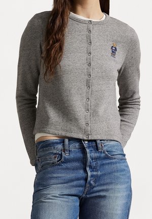 Kvinde iført en grå knapfast cardigan med et lille broderet bjørnelogo på brystet og blå jeans med synlige lommer.