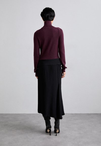 Burgundijas turtlenecks džemperis, apvienots ar melnu midi svārku, kas piedāvā brīvu siluetu, un melniem ceļgaliem augstiem zābakiem ar zelta akcentu.