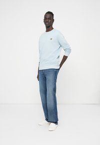 Sweat-shirt bleu clair avec un logo jaune sur la poitrine, associé à un jean bleu et des baskets blanches. Coupe ajustée, design décontracté.