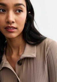 Camisa de color marrón con cuello, hecha de tejido texturizado con rayas verticales y cierre de botón, llevada por una persona con cabello largo y negro.