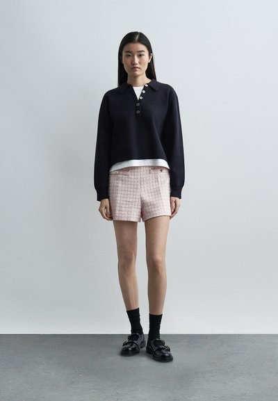 Νεαρή γυναίκα όρθια, φοράει μαύρο πουλόβερ με μακριά μανίκια, λευκό εσώρουχο, ανοιχτό ροζ υφασμάτινες σορτς, μαύρες κάλτσες και μαύρα μοκασίνια.