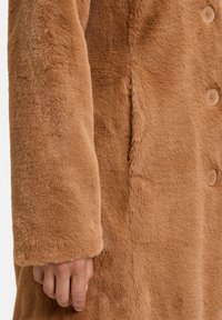 Faux fur frakke i lys brun med en blød tekstur. Har store knapper og en afslappet pasform. Ærmerne er let manchetteret.