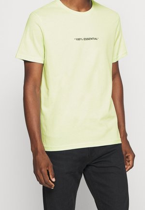 Mężczyzna w jasnżółtej koszulce z krótkim rękawem z napisem „100% ESSENTIAL” na piersi i czarnych spodniach na jednolitym tle.