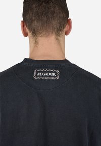 Juodas trumpas rankovės marškinėliai su apvalia kaklo iškirpte ir išsiuvinėtu "PEGADOR" logotipu ant stačiakampio pleistro nugaros kaklo srityje.