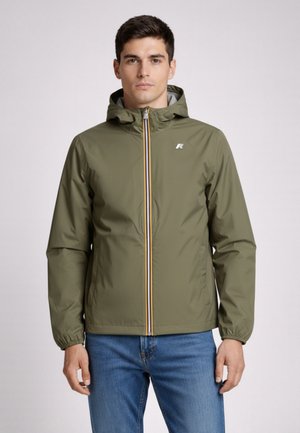 Uomo con giacca con cappuccio verde oliva e cerniera frontale, e jeans blu, in piedi contro uno sfondo chiaro uniforme.