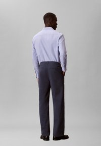 Άνδρας που στέκεται με την πλάτη, φορώντας γαλάζιο πουκάμισο, σκούρο μπλε παντελόνι και μαύρα παπούτσια, μπροστά σε απλό γκρι φόντο.