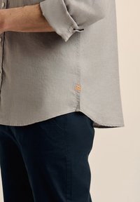 Camisa gris claro con cuello abotonado, mangas largas y un detalle bordado en naranja en el dobladillo, combinada con pantalones azul marino.
