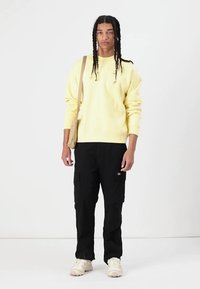 Yellow sweatshirt, black cargo pants, beige shoulder bag, wearing light-colored sneakers, standing against a white background.