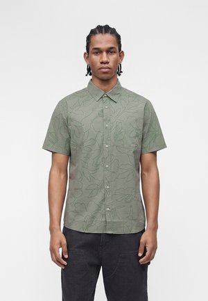 Νεαρός άνδρας με πλεξούδες, που φοράει πράσινο πουκάμισο με κοντά μανίκια και διακριτικό λουλουδένιο σχέδιο και μαύρο παντελόνι σε λευκό φόντο.