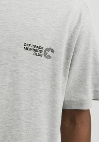 Camiseta de manga corta gris hecha de una tela suave, con un logo impreso en negro que dice "OFF-TRACK MEMBERS’ CLUB" en la parte superior izquierda.