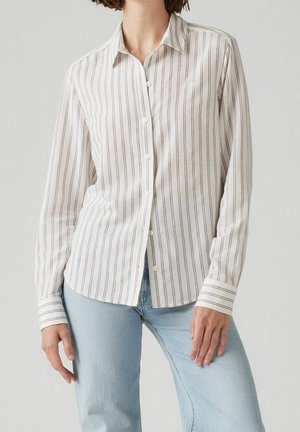Femme portant une chemise blanche à manches longues boutonnée avec de fines rayures noires verticales et un jean bleu clair.