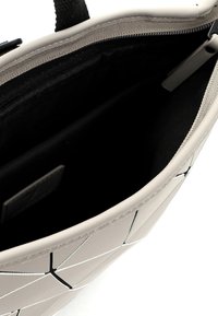 Sac à main beige avec un design à panneaux géométriques, partiellement ouvert pour montrer l'intérieur en tissu noir et une fermeture éclair.