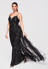 Femme en robe noire ajustée à sequins avec fines bretelles, décolleté profond et longue traîne plissée transparente, debout devant un fond clair uni.