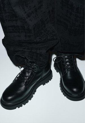 Czarne skórzane botki na sznurowadła z grubą, wytrzymałą podeszwą noszone z luźnymi czarnymi teksturowanymi spodniami na białym tle.