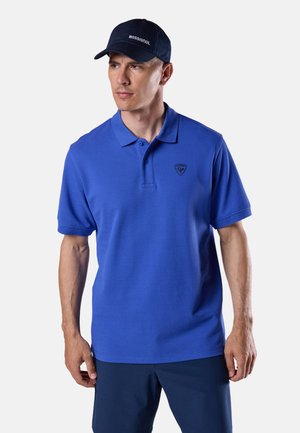 Mann trägt ein blaues Poloshirt und eine marineblaue Kappe mit dem "Rossignol"-Logo, steht vor einem einfarbigen hellen Hintergrund.