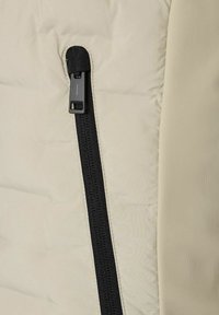 Beige isolierte Jacke mit einem schwarzen Reißverschluss, der ein gestepptes Muster aufweist und eine glatte Textur im Bereich um den Reißverschluss hat.