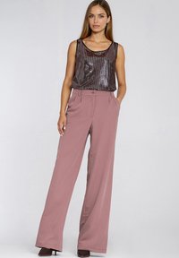 Kobieta w bezrękawniku o metalicznym, ciemnym kolorze i wysokich, szerokich, brudnoróżowych spodniach z rękami w kieszeniach na jednolitym tle.