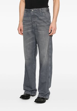 Persona che indossa jeans grigi larghi in denim e scarpe nere in piedi su uno sfondo bianco.