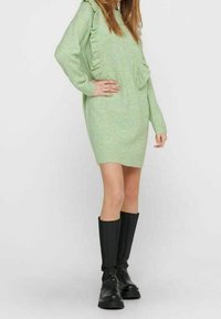 Zielona dzianinowa sukienka z kapturem, ozdobiona falbankami po bokach. Połączona z czarnymi kozakami do kolan. Gładka faktura.