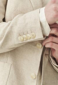Bež jakna od lana sa rukavima koji sadrže četiri kreme boje dugmeta, od kojih svako ima dvije rupe, te jedno dugme na manžetama. Vidljiva teksturirana tkanina.
