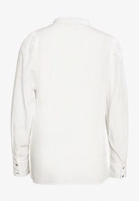 Blusa bianca a maniche lunghe con tessuto strutturato, colletto arricciato e polsini con bottoni. Presenta una schiena liscia senza motivi.
