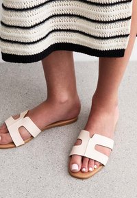 Beige leren slip-on sandalen met uitsnijdingen en een gladde zool. Een voet met een frisse pedicure is gedeeltelijk zichtbaar, geplaatst tegen een gestructureerde vloer.