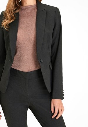Femme portant un blazer noir cintré par-dessus un top marron texturé et un pantalon noir assorti, posant avec une main relâchée sur le côté.