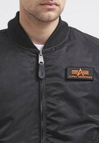 Jachetă bomber neagră cu guler ribbed, fermoar în față și patch cu logo din material portocaliu. Material neted cu un ușor luciu. Design ajustat.