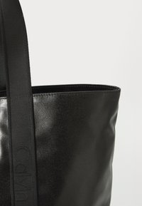 Saco tote preto feito de material sintético brilhante, apresentando uma alça larga preta com logótipos em relevo e um design de topo aberto.