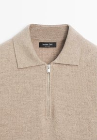 Beżowy sweter z dzianiny z kołnierzem na zamek, o miękkiej fakturze i subtelnym wzorze. Metka marki widoczna przy dekolcie.