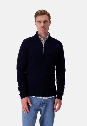 Marineblauer, gestrickter Pullover mit einem Viertel-Reißverschluss-Kragen und einer Fronttasche; über einem hellblauen Hemd mit Knöpfen getragen und mit hellen Jeans kombiniert.