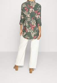 Groene bloemenblouse met driekwartmouwen, versierd met roze bloemen en vlinders, gecombineerd met wijde witte broeken en bruine blokhakken.