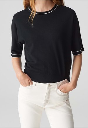 Femme portant un pull noir à manches courtes avec des manches bordées de blanc et un jean taille haute écru, les mains dans les poches, sur un fond uni.