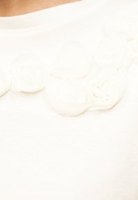 Κοντινό πλάνο λευκού υφάσματος με λεπτά φλοράλ δαντελένια κοσμήματα κατά μήκος μιας καμπύλης γραμμής λαιμού.