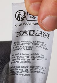 Etichetta di cura con tessuto bianco che mostra simboli di lavanderia e testo in più lingue, dettagliando la composizione del materiale e le istruzioni di lavaggio.