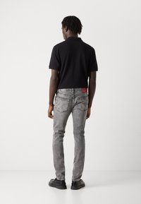 Camisa polo negra de manga corta, combinada con jeans gris desgastados y zapatos negros, vista desde la parte trasera. Etiqueta roja visible en los jeans.