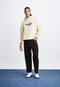 Beige capuchontrui met grafische print, gecombineerd met donkerbruine corduroybroek en witte sneakers. Soepele stof en relaxte pasvorm.