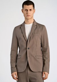 Blazer marrón de tela texturizada con dos bolsillos frontales, cierre de un solo botón, solapas con muesca y corte ajustado, llevado sobre una camiseta blanca.