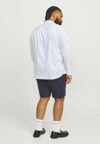 White long-sleeve shirt with button cuffs, worn with navy shorts. Model stands on a plain background, showcasing the outfit from the back.