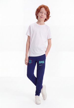 Enfant aux cheveux roux debout avec une main dans la poche, portant un t-shirt blanc, un pantalon bleu avec les logos Barça et Nike, et des baskets blanches sur fond blanc.
