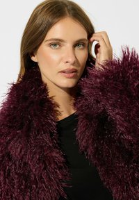 Lungo cappotto peloso di un profondo colore bordeaux, con texture shaggy e un ampio colletto. Indossato sopra una camicia nera, con focus su viso e spalle.