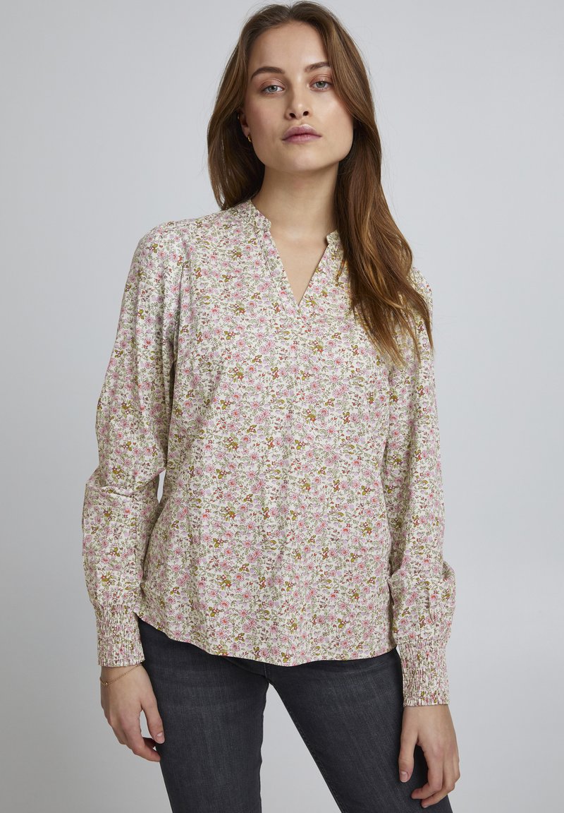 Blomstret bluse i let stof med V-udskæring. Har et pink, grønt og hvidt blomstermønster, lange ærmer og sammentrukne manchetter.