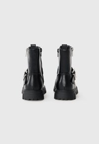 Bottines en cuir noir avec une semelle épaisse, des zips à l'arrière et des boucles métalliques décoratives sur le côté. Texture lisse et design moderne.