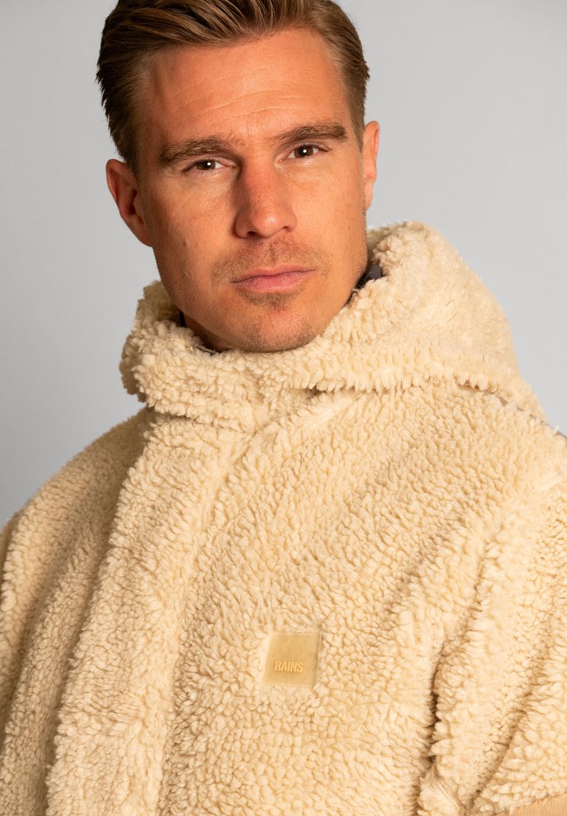 Uomo con capelli biondi corti che indossa un giacca di tessuto soffice e color crema con colletto alto e un'etichetta "RAINS" sul petto, su uno sfondo neutro.