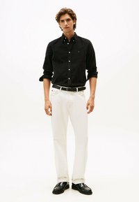 Zwarte button-up shirt, witte jeans, zwarte gevlochten riem, zwarte schoenen, staand tegen een effen witte achtergrond. Details van de casual outfit zichtbaar.