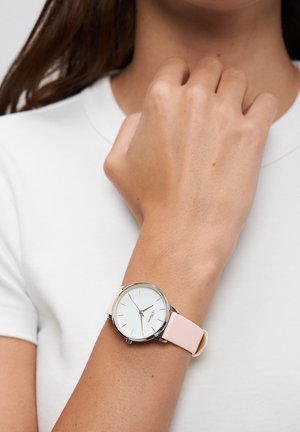 Round silver watch with a white dial, black hour markers, and a light pink silicone strap. Worn on a wrist with natural skin texture.